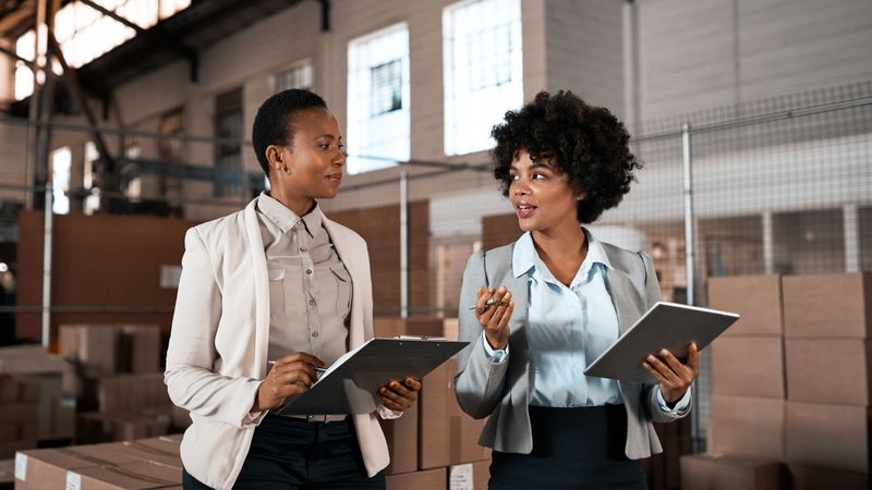 Two commercial operations leads reviewing records on a clipboard and tablet in a distribution warehouse